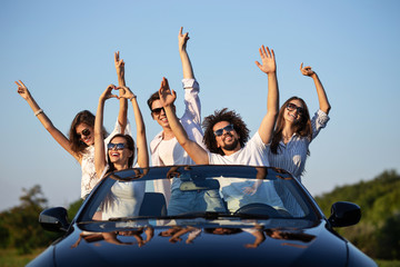 Stylish young girls and guys in sunglasses are sitting in a black cabriolet on the road holding...