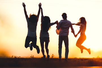 Two young guys and two girls are holding their hand and jumping in the field on a summer day. Back view.