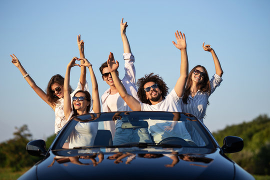 Stylish young girls and guys in sunglasses are sitting in a black cabriolet on the road holding their hands up and smiling on a sunny day.