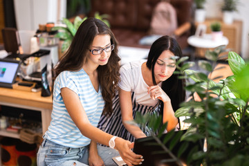 Two good looking young girls with long dark hair,wearing casual style,sit at the table and look attentively at the laptop screen in a cozy coffee shop.