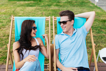 A beautiful couple in the sunglasses is lying on the deck chairs on the lawn in the nice summer cafe. Entertainment, having good time. Friendship, relationship.