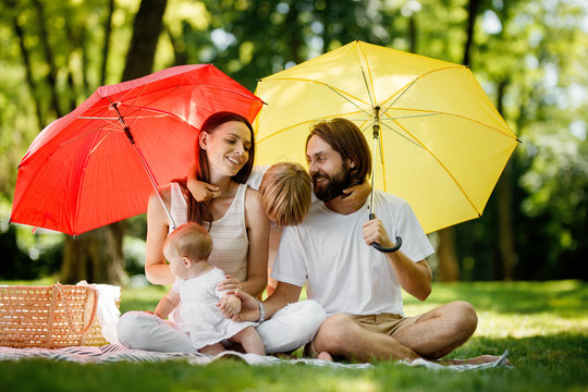 brunettes parents with two kids have a rest on the lawn under the bright red and yellow umbrellas covering them from the sun.