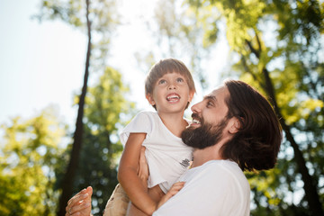 Fototapeta premium Handsome dark-haired father with beard dressed in the white t-shirt is holding in the arms his little son, watching at him and smiling in the park.