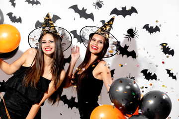 Two charming young women in witches hats hold black and orange balloons on a white background with black bats. Confetti around