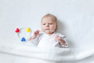 Cute adorable newborn baby playing with colorful rattle toy. in white bed at home. New born child, little girl looking surprised at the camera. Family, new life, childhood, beginning concept.