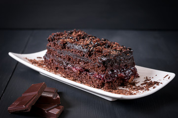 Pie with layers of cream, sprinkled with grated chocolate, on a white saucer on a black wooden background	