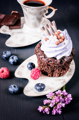 Chocolate cupcake with icing on a saucer with berries and coffee cup on a black wooden background	