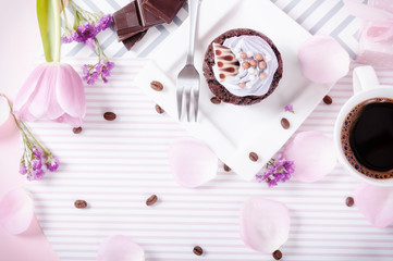 Black coffee and chocolate cupcake with icing on a pink background with tulip petals and coffee beans