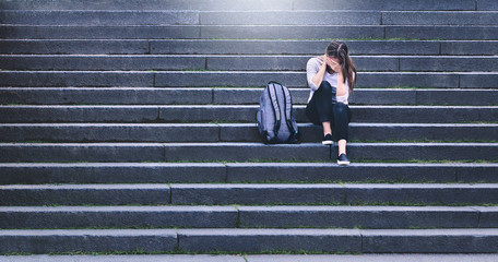 Bullying, discrimination or stress concept. Sad teenager crying in school yard. Upset young female student having anxiety. Upset victim of abuse or harassment sitting on stairs outdoors with backbag.