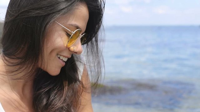 Lovely shy caucasian woman in stylish sunglasses cutely smiles straight to camera on sea background. Summer vacation, chilling, lighten up. Female portrait, close up view