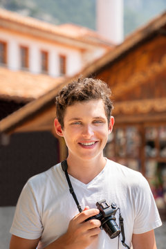 Portrait of a young teenage male holding a camera outdoors smiling. Blurred buildings in the background.