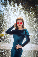 Portrait of beautiful young woman with bright makeup, wearing emerald green velvet dress and red sunglasses, standing near fountain at city street