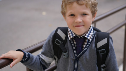 Little Caucasian curly boy in school uniform with backpack comes up on stairs.