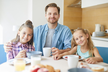 Young father and his two adorable daughters looking at you while sitting by served table in the kitchen