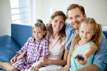 Young cheerful family of four sitting on sofa in front of tv set while one of them switching...
