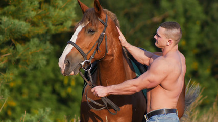 Obraz premium Young strong guy standing next to a horse in the field.