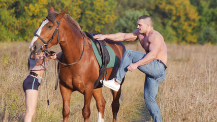 Young man with a strong physique climbs on a horse, a young slender woman holding a horse