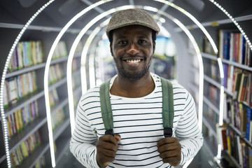 Smiling guy in striped long-sleeved vest with backpack behind his back standing in college library