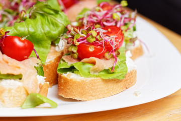 Fresh homemade small sandwiches with sprouts, smoked salmon, bread, lettuce, smoked salmon, coctail tomato, and white cheese on a wooden table prepared to house party