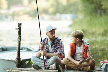 Young man with rod and his son sitting on wooden pontoon and talking while fishing on countryside