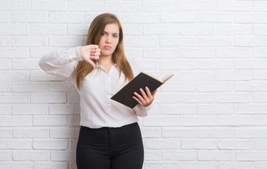 Young adult woman standing over white brick wall reading a book with angry face, negative sign showing dislike with thumbs down, rejection concept