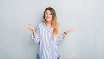 Young adult woman over grey grunge wall wearing fashion business outfit clueless and confused expression with arms and hands raised. Doubt concept.