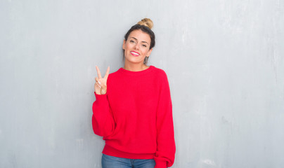 Obraz na płótnie Canvas Young adult woman over grey grunge wall wearing winter outfit smiling with happy face winking at the camera doing victory sign. Number two.
