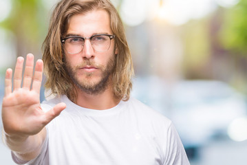 Young handsome man with long hair wearing glasses over isolated background doing stop sing with palm of the hand. Warning expression with negative and serious gesture on the face.