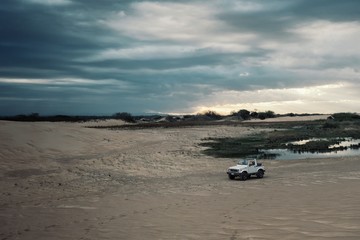 4x4 car waiting next to a lagoon in front of the sand dunes at sunset with beautiful sky