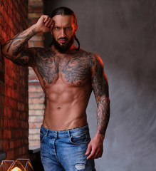 Fototapeta na wymiar Pensive shirtless bearded male with the muscular and tattooed body leaning on a wall and looks at the camera.