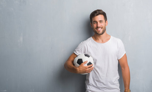 Handsome young man over grey grunge wall holding soccer football ball with a happy face standing and smiling with a confident smile showing teeth
