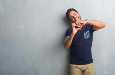Handsome young man over grey grunge wall smiling in love showing heart symbol and shape with hands. Romantic concept.