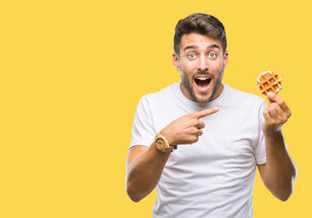 Young handsome man eating a sweet waffle over isolated background very happy pointing with hand and...