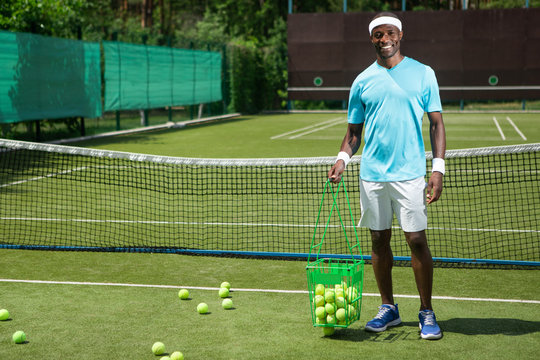 Full length portrait of grinning athlete standing on tennis playground on sunny day. He is holding basket with balls while some of them are lying on ground. Copy space in left side