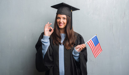Young brunette woman over grunge grey wall wearing graduate uniform holding flag of America doing ok sign with fingers, excellent symbol