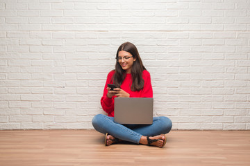 Young brunette woman sitting on the floor using laptop and smartphone with a happy face standing and smiling with a confident smile showing teeth