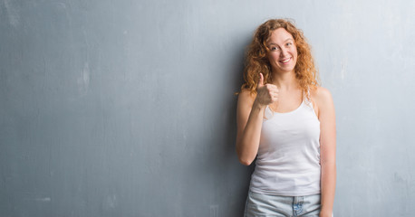 Fototapeta premium Young redhead woman over grey grunge wall happy with big smile doing ok sign, thumb up with fingers, excellent sign