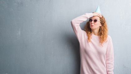 Young redhead woman over grey grunge wall wearing birthday cap stressed with hand on head, shocked with shame and surprise face, angry and frustrated. Fear and upset for mistake.