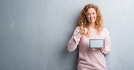 Young redhead woman over grey grunge wall using tablet happy with big smile doing ok sign, thumb up with fingers, excellent sign