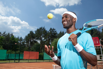 Smiling sportsman is playing match in sun. He is holding racket and throwing up ball with hand. Copy space in left side