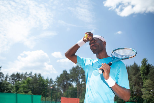 Low angle of man standing and holding racket and ball. He is lifting hand and closing face from light while pursing lips. Copy space in left side