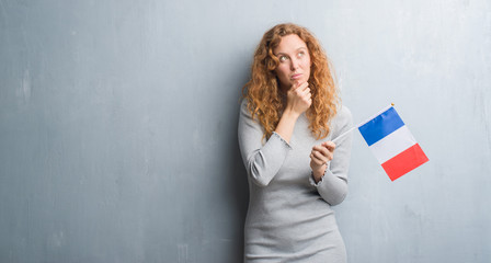 Young redhead woman over grey grunge wall holding flag of France serious face thinking about question, very confused idea