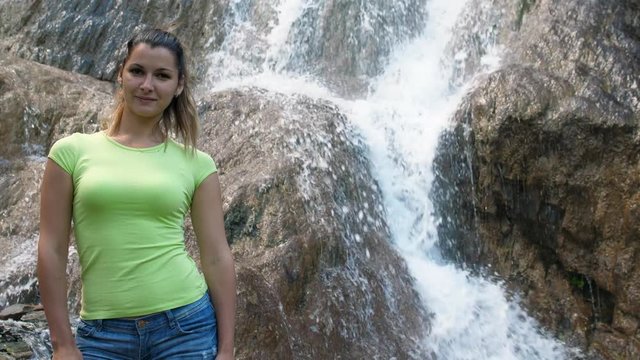 A woman stands on a background of a waterfall. Rapid waterfall running down the rock. Beautiful smiling girl next to a stream of water.
