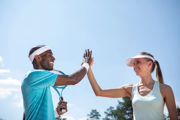 Tuinposter Low angle of positive couple playing tennis outdoor on warm sunny day. They are high-fiving while celebrating victory after tennis match. Athletes are standing beside while guy is holding racket © Yakobchuk Olena