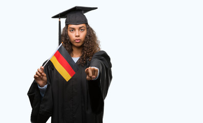 Young hispanic woman wearing graduation uniform holding flag of Germany pointing with finger to the camera and to you, hand sign, positive and confident gesture from the front