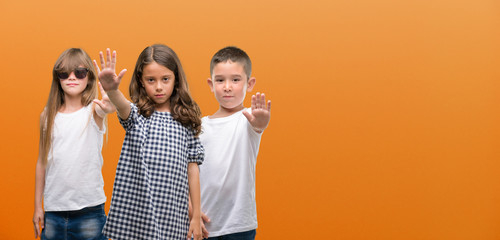 Group of boy and girls kids over orange background with open hand doing stop sign with serious and...