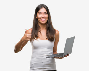 Young hispanic woman using computer laptop happy with big smile doing ok sign, thumb up with fingers, excellent sign