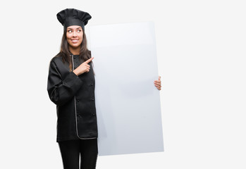 Young hispanic cook woman wearing chef uniform holding banner very happy pointing with hand and finger to the side