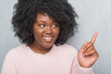 Young african american plus size woman over grey grunge wall wearing winter sweater with a big smile on face, pointing with hand and finger to the side looking at the camera.