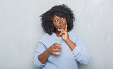 Young african american woman over grey grunge wall drinking a glass of water serious face thinking...